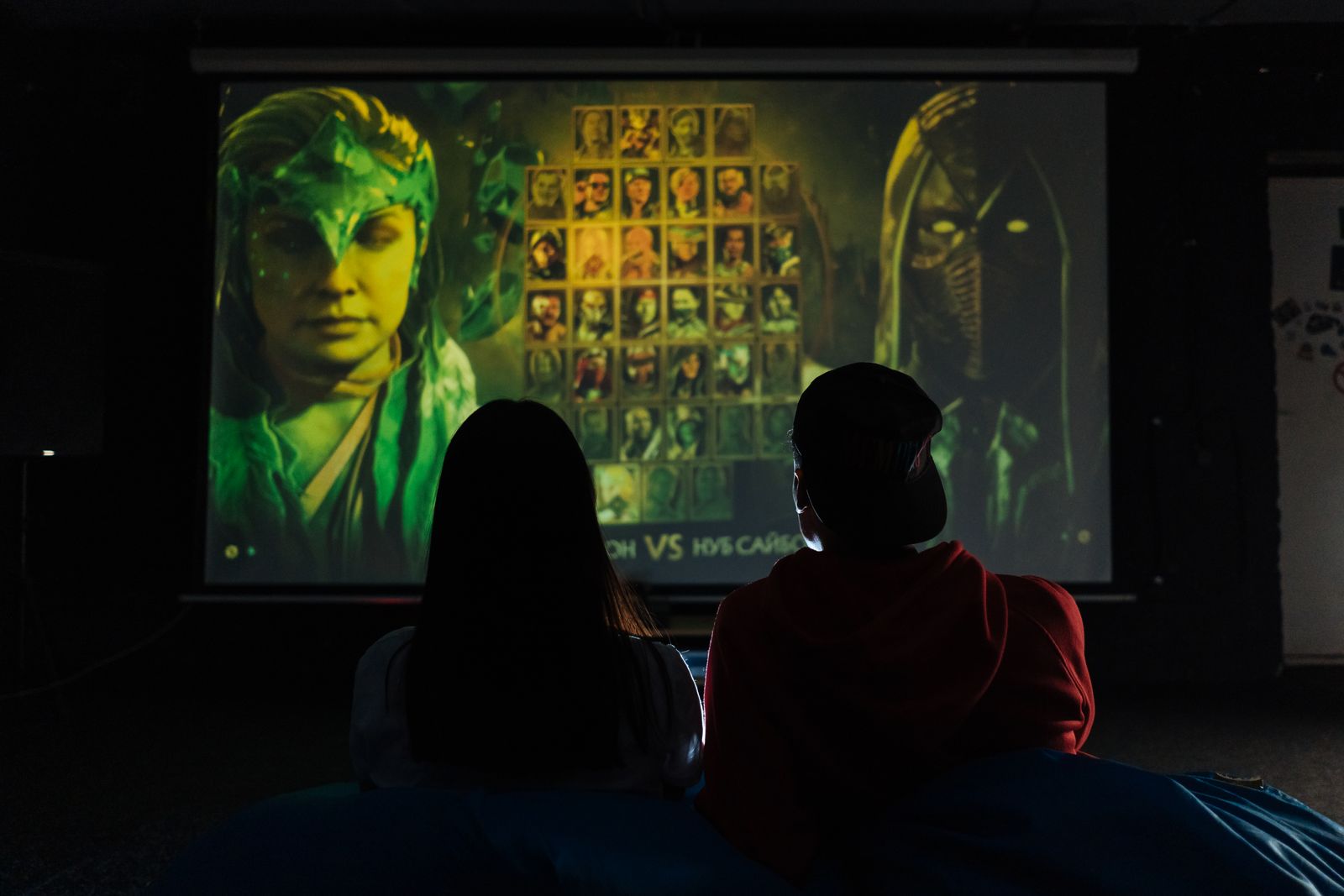 Two people gaming indoors on a large projector screen in a dim room.