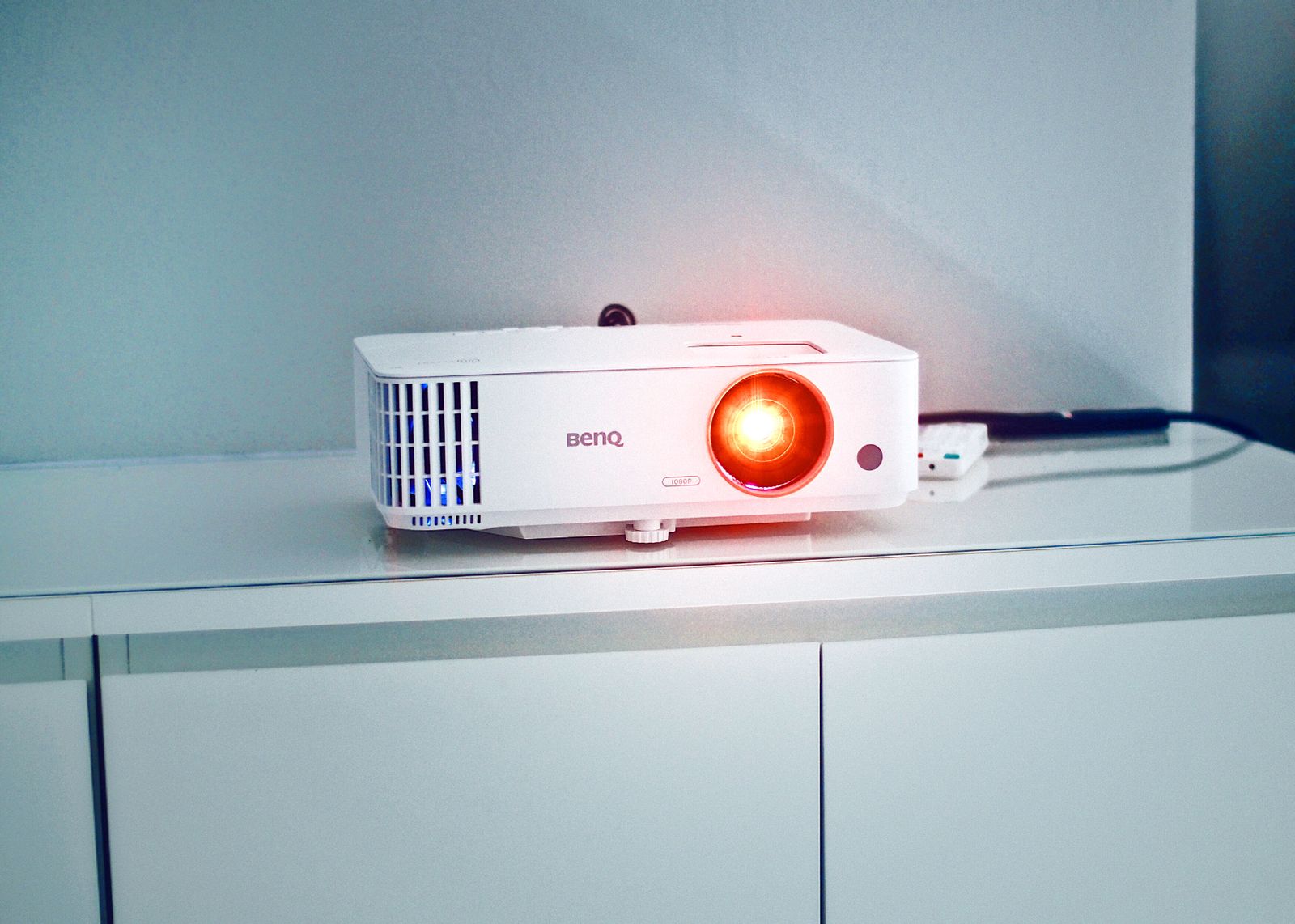 A white projector with a glowing lens sitting on a cabinet
