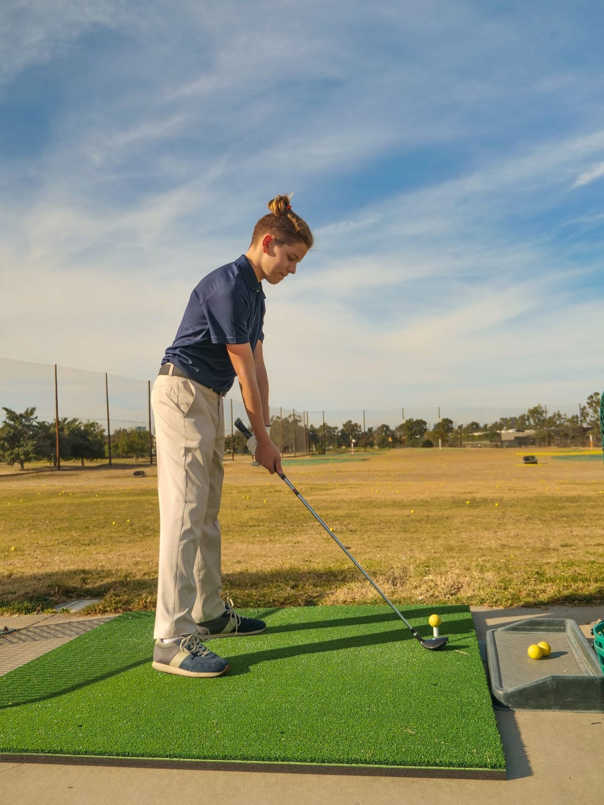 A golfer in a navy polo at address over a synthetic hitting mat, a yellow ball teed up with the clubhead set behind it.