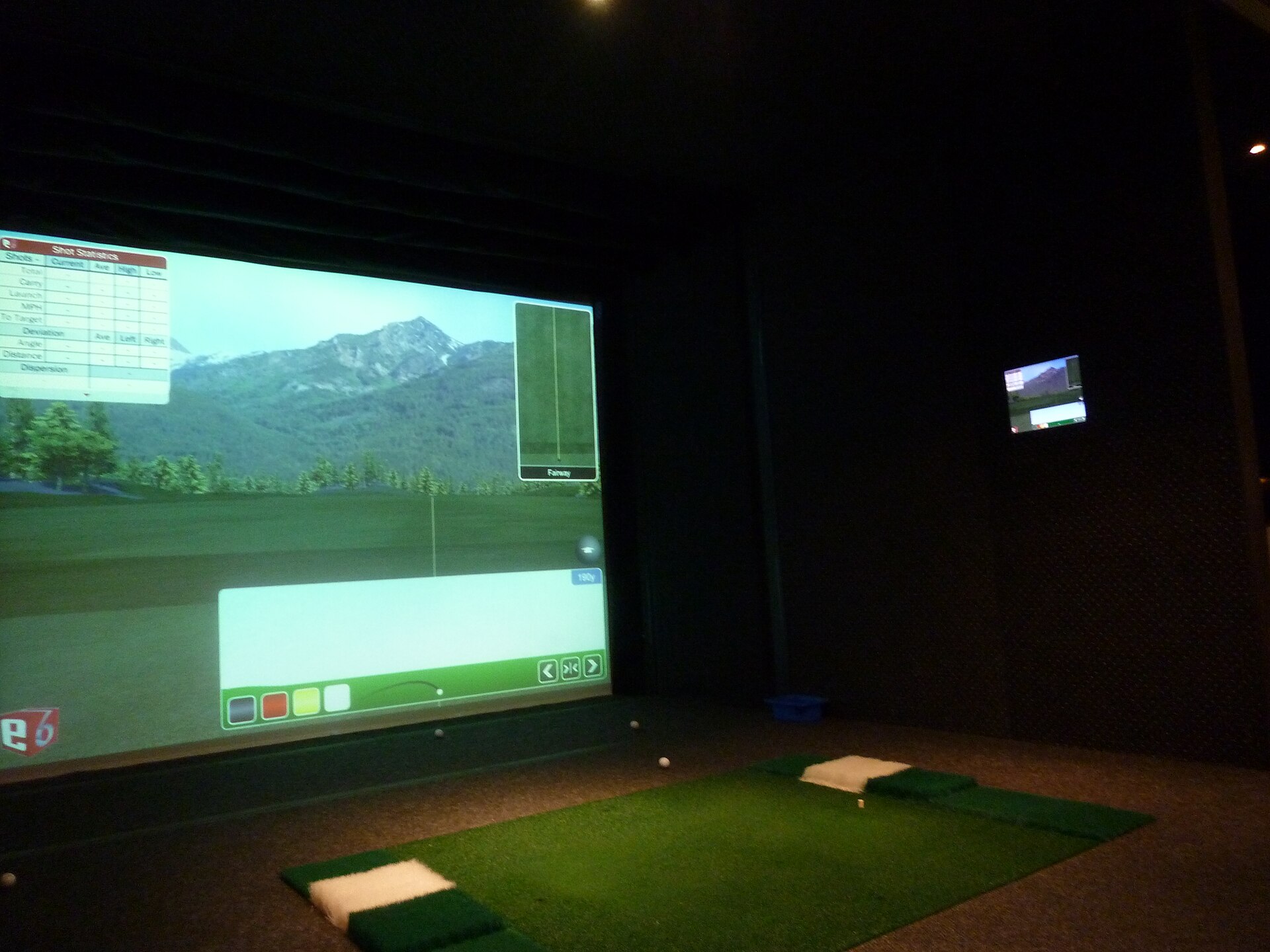 An indoor golf simulator bay with a projected course on the impact screen, a shot-statistics overlay and a hitting mat