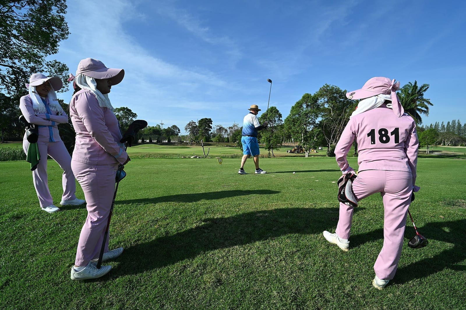 Three caddies in matching uniforms and wide-brimmed sun hats stand on a tee holding covered clubs, watching a golfer play his shot down a sunlit fairway.